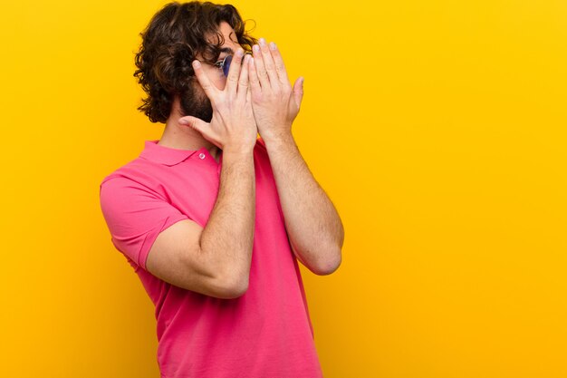 Young crazy man covering face with hands, peeking between fingers with surprised expression and looking to the side  orange wall