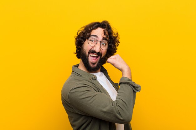 Young crazy man feeling happy, satisfied and powerful, flexing fit and muscular biceps, looking strong after the gym  yellow wall