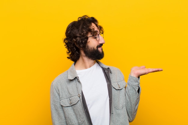 Young crazy man feeling happy and smiling casually, looking to an object or  held on the hand on the side  yellow wall