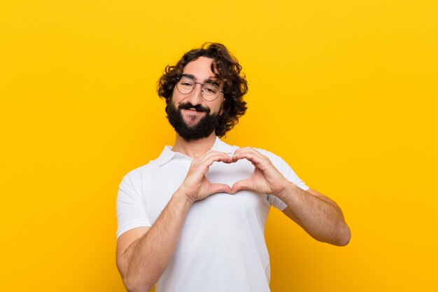 Young crazy man smiling and feeling happy, cute, romantic and in love, making heart shape with both hands against yellow wall