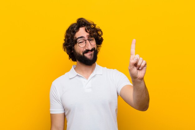 Young crazy man smiling and looking friendly, showing number onefirst with hand forward, counting down  yellow wall