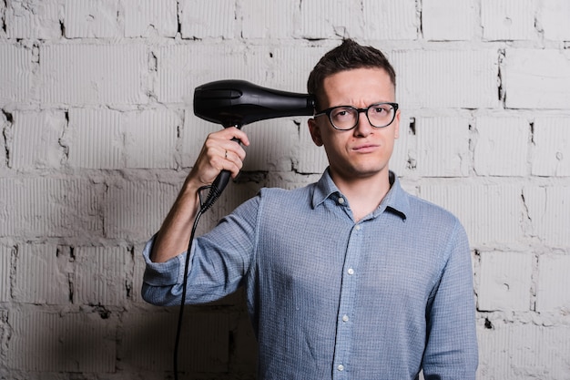 Young crazy stylish man with hairdraier and funny expressions on grey brick wall