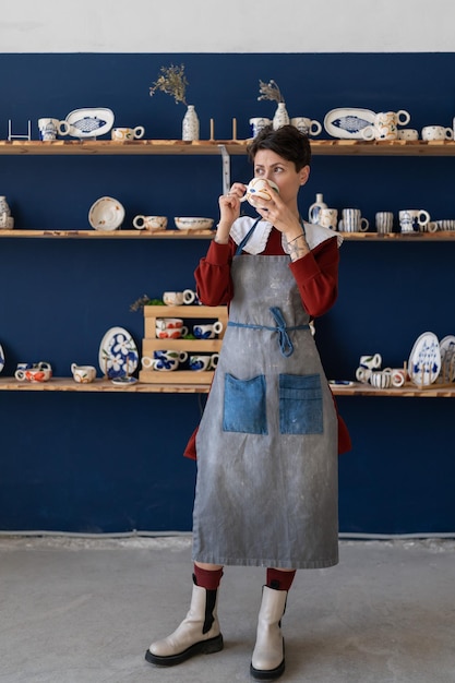 Young creative artisan crafts woman wearing apron drinking\
morning coffee from handmade clay mug