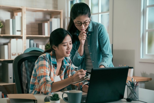 Young creative business people at office. two asian woman colleague in bright vintage studio talking on festival event plan on notebook and laptop pc against window. female coworker teach intern