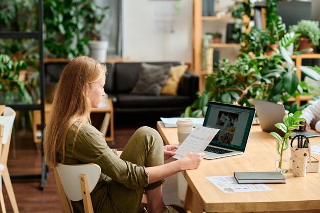 Young creative businesswoman or designer with document looking at laptop screen