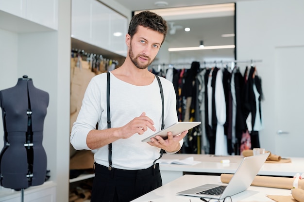 Young creative fashion designer with touchpad looking at you while browsing in the net for new models for collection