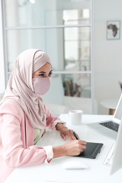 Young creative female designer in hijab and protective mask sitting by desk in front of computer and retouching images or drawing sketch