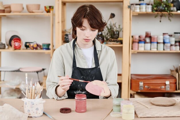 Photo young creative female in workwear painting handmade earthenware