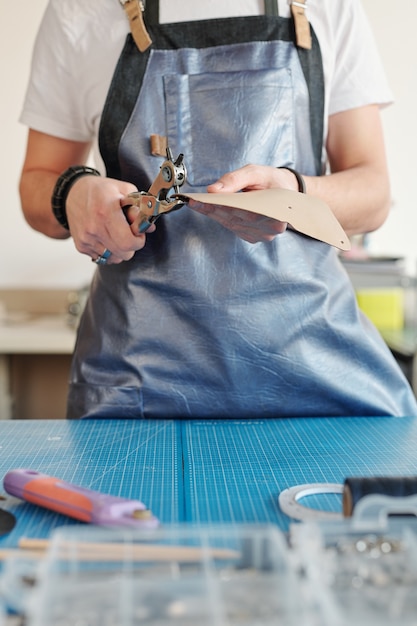 軽い革の部分を保持し、穴を開けながらテーブルのそばに立っている作業服の若い創造的な男