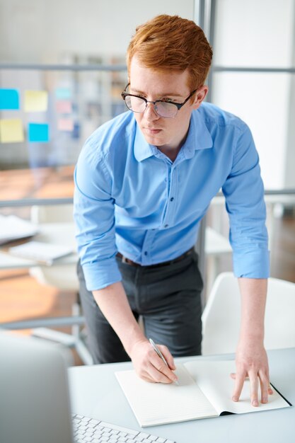 Young creative student or designer making notes in copybook while looking at computer screen