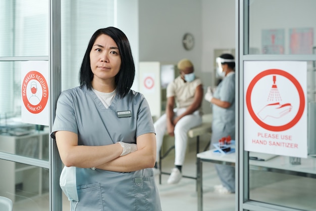 Young cross-armed female clinician in uniform standing in front of camera while her colleague vaccinating one of patients in medical office