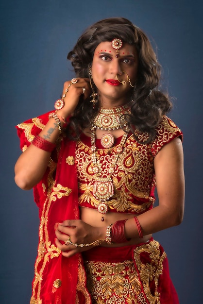 Young crossdresser man wearing Indian wedding clothes of a bride with bridal makeup and posing on grey background looking fashion and glamorous