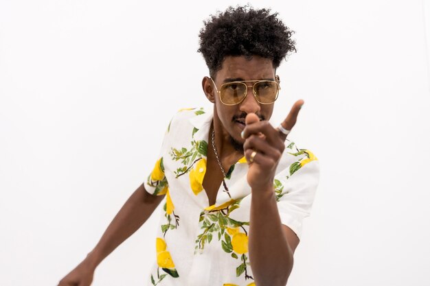 A young Cuban man with curly hair sitting in a white flowered shirt and glasses. Pointing looking at camera on a white background