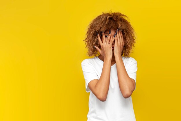 Young curly american girl covers her face with her hands and peeps on yellow isolated background