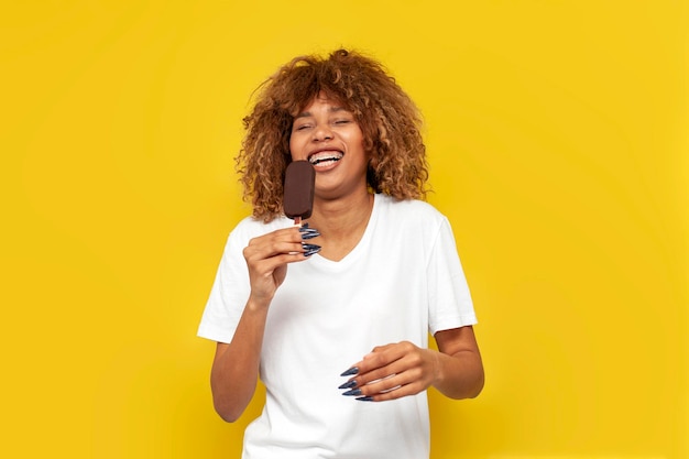 Young curly american girl with braces eats chocolate ice cream on yellow isolated background