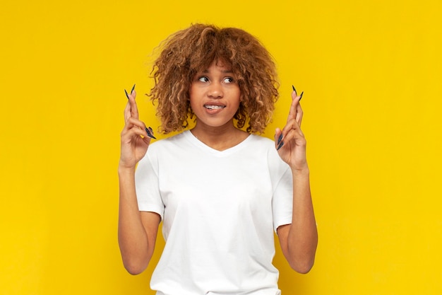 Young curly american girl with braces praying and hoping for good luck on yellow isolated background