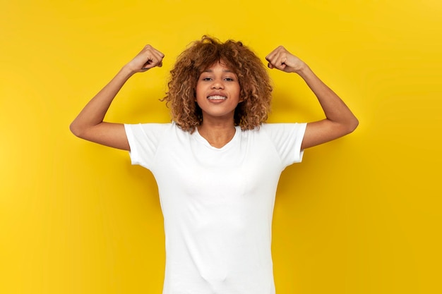 Young curly american girl with braces shows biceps over yellow isolated background