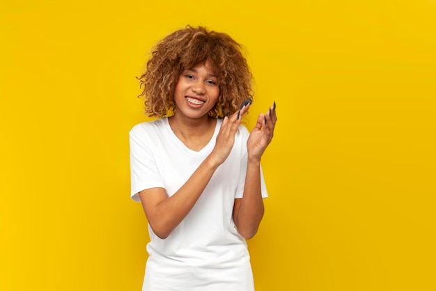 Young curly american girl with braces smiles and applauds on yellow background