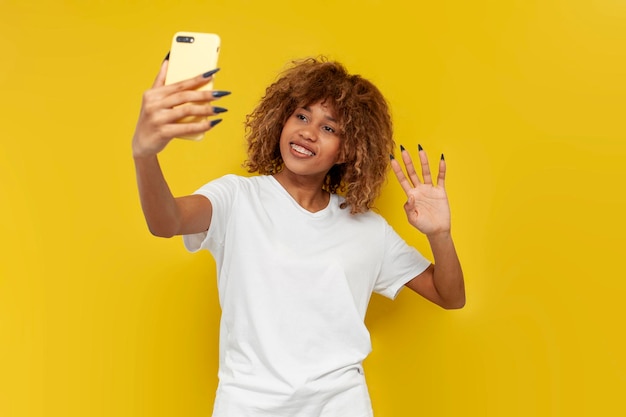 Young curly american girl with braces uses smartphone greets and shows palm on yellow background