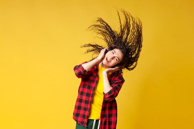 Young curly brunette girl in a plaid shirt listens to music in headphones and dances