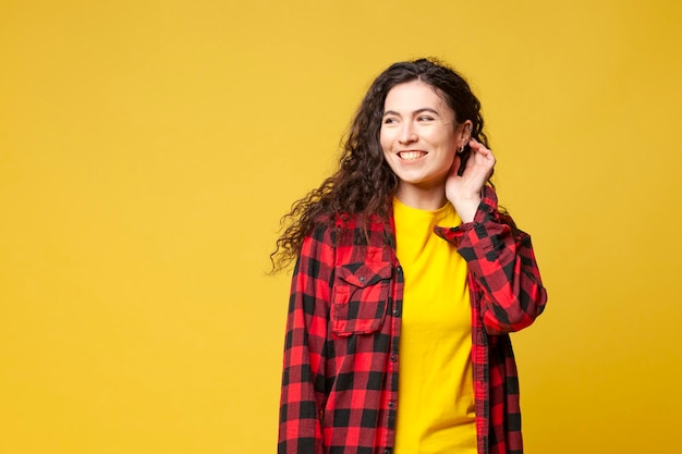 Young curly brunette girl in plaid shirt stands on a yellow background and smiles