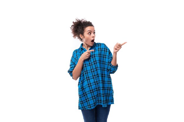 Young curly brunette woman in a blue plaid shirt shows her hand to the side in surprise