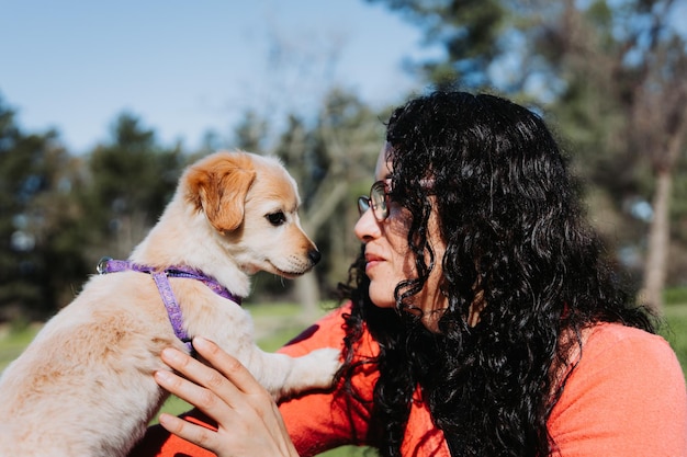 Young curly brunette woman, smiling and playing with a golden retriever puppy in the park.