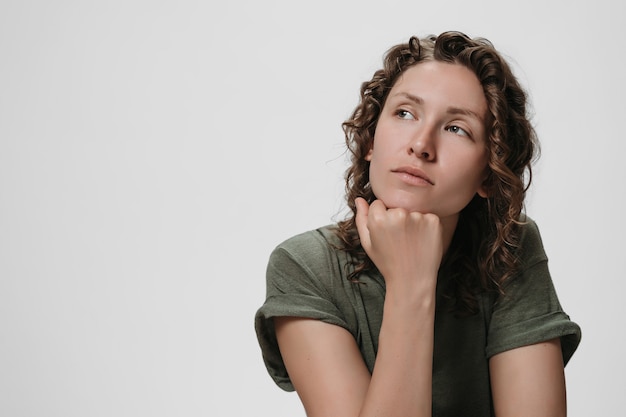 Young curly caucasian woman keeps hand under chin, being deep in thoughts, thinks about something