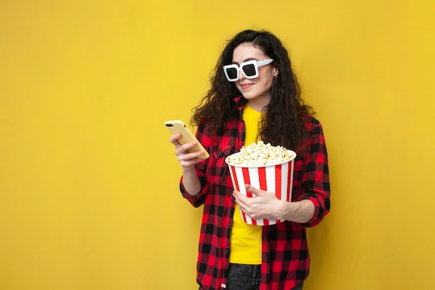 Young curly girl in 3d glasses uses smartphone and holds popcorn on yellow background