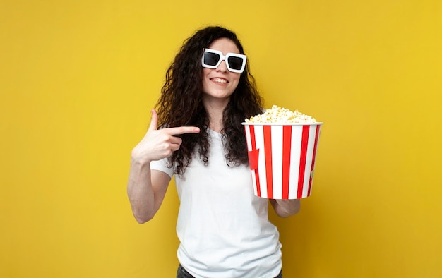 young curly girl in 3d glasses and white tshirt holds popcorn on yellow background