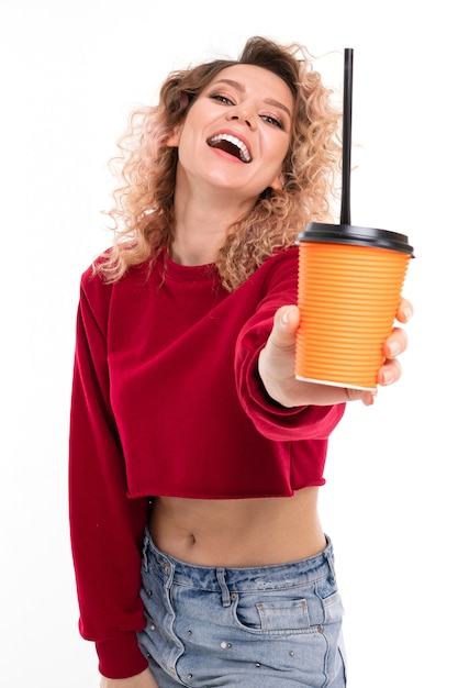 Young curly girl with a smile on her face holds a glass of coffee on outstretched hand