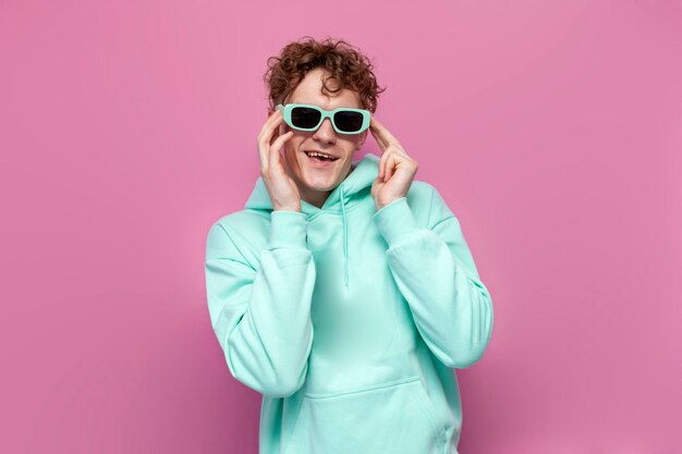 Young curly guy in mint hoodie and sunglasses smiles and dances on pink isolated background