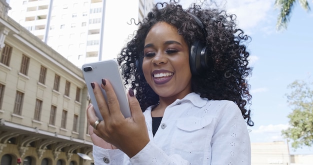 Young curly hair black woman walking using cell phone. Texting on street. Big city.