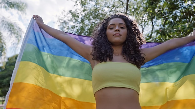 Young curly hair woman covering with lgbt pride flag. Keeping fist up, covering LGBT+ flag