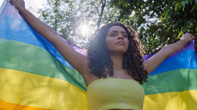 Young curly hair woman covering with lgbt pride flag. Keeping fist up, covering LGBT+ flag