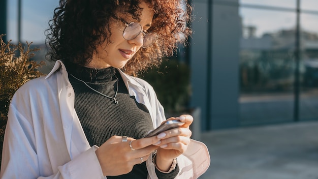 Photo young curly haired freelancer chatting with somebody outside while wearing glasses in a sunny day