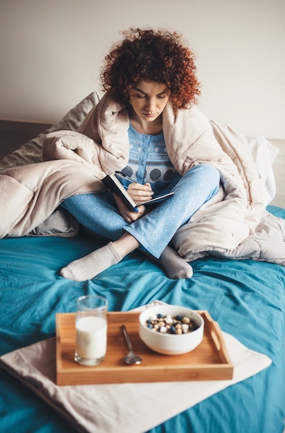 Giovane studente dai capelli ricci scrivendo qualcosa nel libro e mangiando cereali con latte a letto