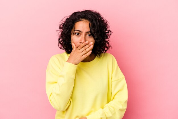 Young curly latin woman isolated on pink background covering mouth with hands looking worried.