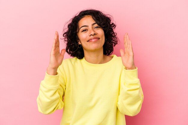 Young curly latin woman isolated on pink background holding something little with forefingers, smiling and confident.