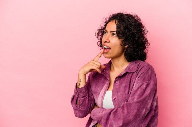 Молодая фигурная латинская женщина, изолированная на розовом фоне, смотрит в сторону с сомнительным и скептическим выражением лица.