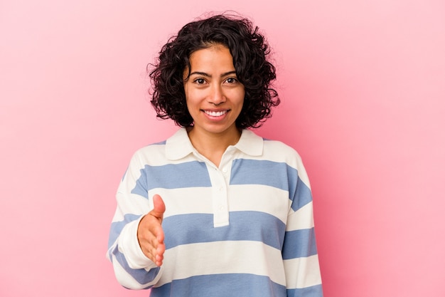 Young curly latin woman isolated on pink background stretching hand at camera in greeting gesture.