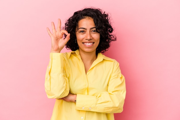 Young curly latin woman isolated on pink background winks an eye and holds an okay gesture with hand.