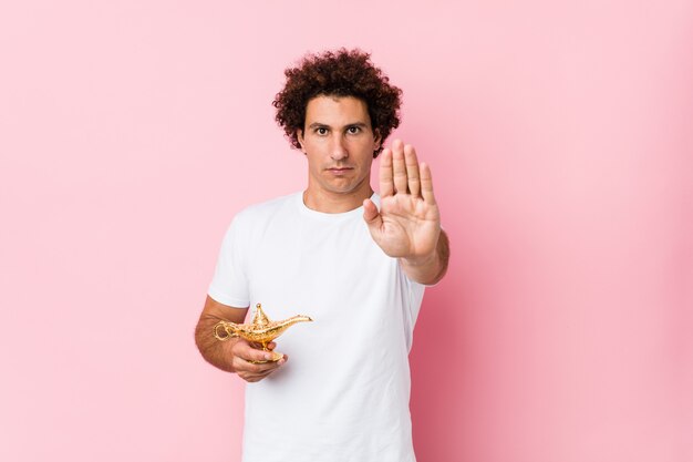 Young curly man  holding a magic lamp standing with outstretched hand showing stop sign, preventing you