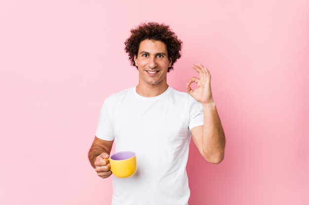 Young curly man holding a tea cup cheerful and confident showing ok gesture