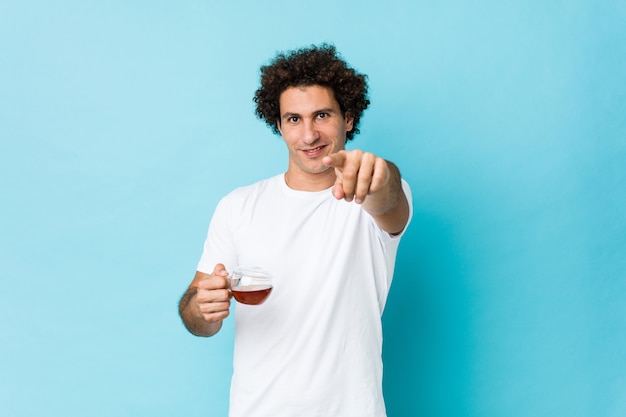 Young curly man holding a tea cup cheerful smiles pointing to front