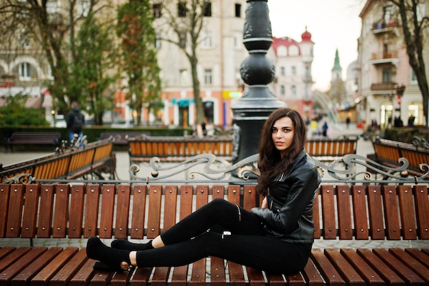 Young curly and sexy woman in leather jacket sitting on bench at street