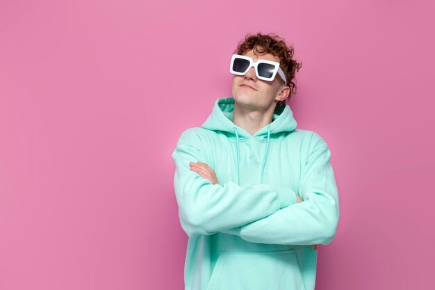 Photo young curly shocked guy in mint hoodie and sunglasses looks away on pink isolated background