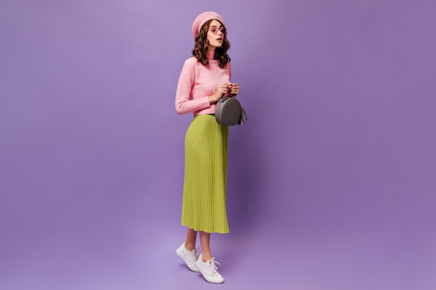 Young curly woman in midi green skirt and pink beret walks on purple wall