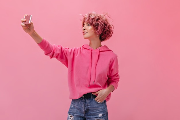 Young curly woman in pink hoodie and denim shorts takes selfie on isolated