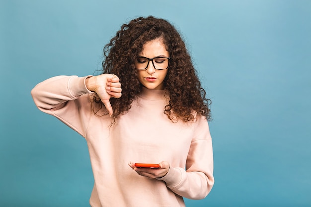 Giovane donna riccia che invia messaggi di testo utilizzando lo smartphone su sfondo blu isolato con la faccia arrabbiata